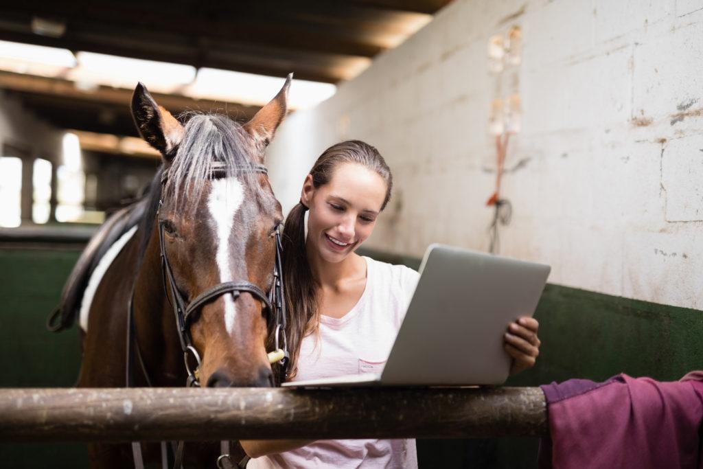 Smiling female jockey using laptop while standing by horse